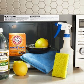 Cleaning supplies arranged in front of a microwave, including vinegar, baking soda, lemons, spray bottle, gloves, sponge, and cloth on a kitchen counter.