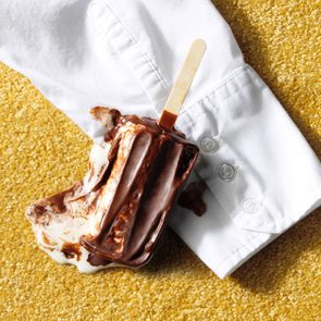 Melted chocolate popsicle on white shirt sleeve atop yellow carpet.