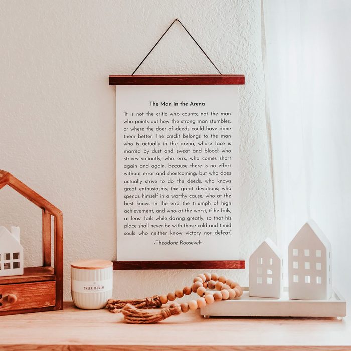 A framed quote hangs on a textured wall, accompanied by small decorative houses and a candle on a wooden shelf.