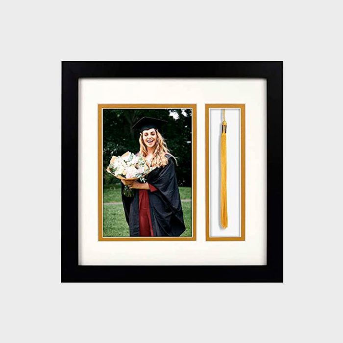 Graduate smiling with bouquet; framed in black, gold-bordered frame alongside a golden tassel, set in an outdoor green park setting.