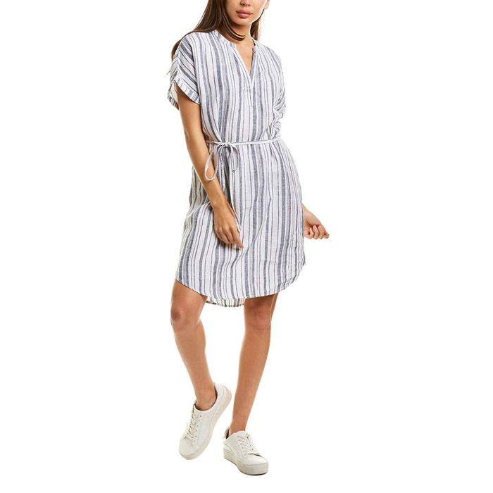 A person stands wearing a short-sleeve, striped dress with a waist tie in a white background, paired with white sneakers.