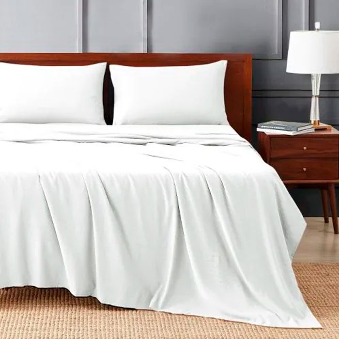 A bed with white sheets and pillows sits in a room with a wooden nightstand and a lamp, on a woven rug.