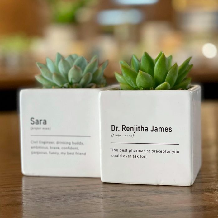 Succulent plants sit in square, white pots with personalized texts, placed on a wooden surface. Background features blurred indoor lights.