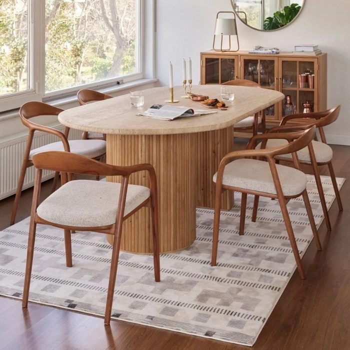 Wooden dining table displays open magazines and glasses, surrounded by modern chairs, in a bright room with large windows and a patterned rug.