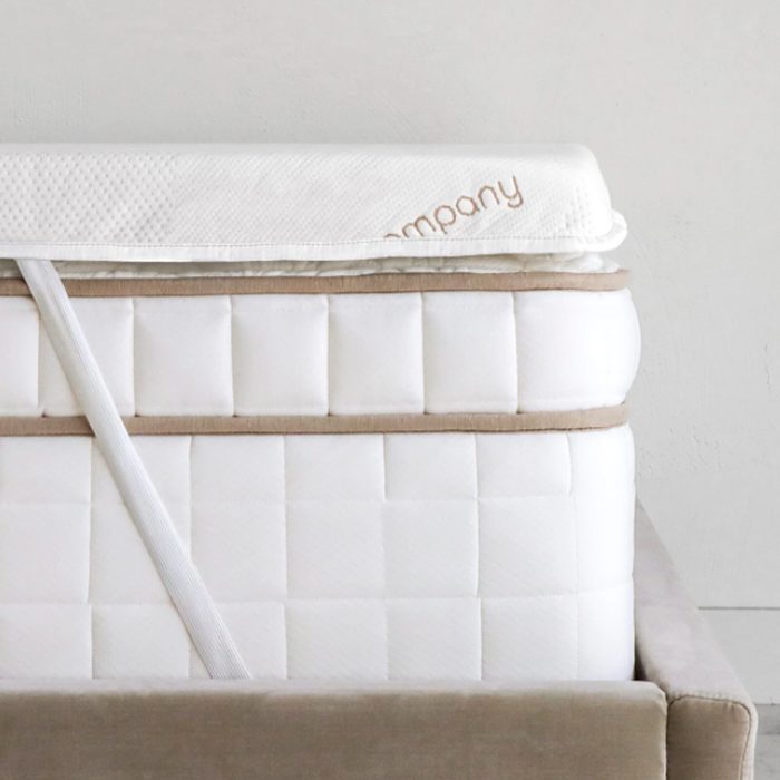 Mattress stack includes a white topper and thick mattress, secured with a strap, against a gray wall backdrop. Text reads 