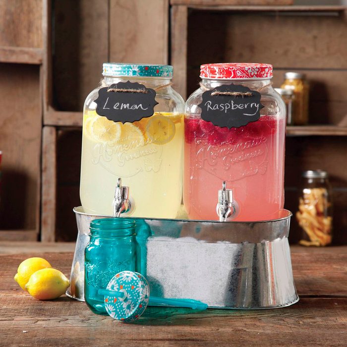 Jars of lemon and raspberry drinks sit with spouts in a metal tray, rustic background, beside lemons and a blue jar on a wooden surface.