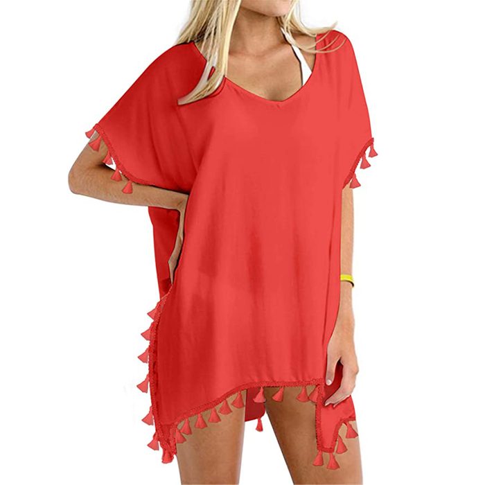 A person wears a red cover-up with tassels, over a swimsuit, standing casually in a bright, neutral setting.