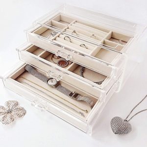 Jewelry box displays rings, watches, and necklaces in clear drawers, set on a white surface with additional jewelry alongside.