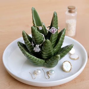 A ceramic cactus holds rings, surrounded by earrings and small jewelry, on a white dish; a corked bottle is in the background.