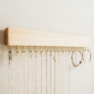 Wooden rack holding necklaces and hoop earrings, hanging neatly from small hooks against a plain light-colored wall.