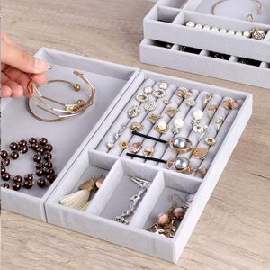 Jewelry is neatly organized in a grey velvet tray on a wooden surface, including bracelets, earrings, and rings. A hand holds one bracelet.