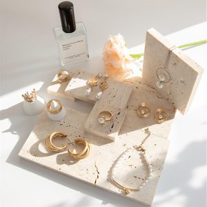 Jewelry displayed on textured stone platforms; includes earrings, rings, and a necklace. Nearby, a perfume bottle and a peach flower provide elegant context.