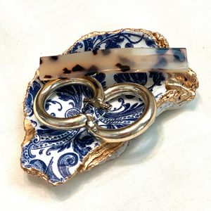 Decorative dish holds two hoop earrings and a tortoiseshell barrette, featuring a blue floral pattern and gold edges on a white surface.