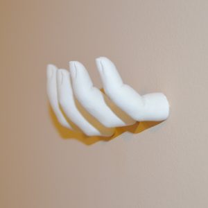 White ceramic hand extends horizontally, palm facing up, against a plain beige wall. Shadows form beneath the fingers, enhancing the three-dimensional effect.