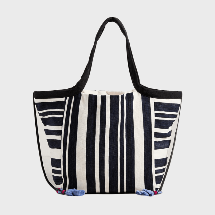 Striped tote bag with black handles sits against a plain gray background, featuring vertical navy and white stripes with small blue tassels.
