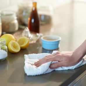 Hand wipes kitchen counter with cloth, surrounded by lemons, a jar of baking soda, a blue container, and other kitchen items.