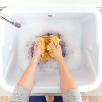 Hands wash yellow fabric in soapy water within a white basin on a light countertop.