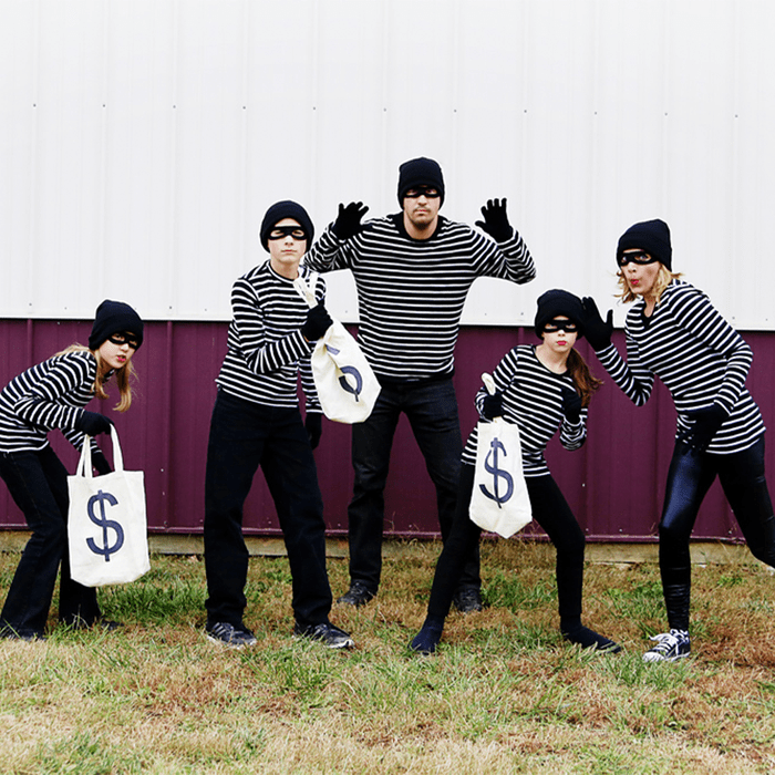 Five people, dressed as burglars with striped shirts and masks, crouch with dollar bags outside on grass in front of a building.