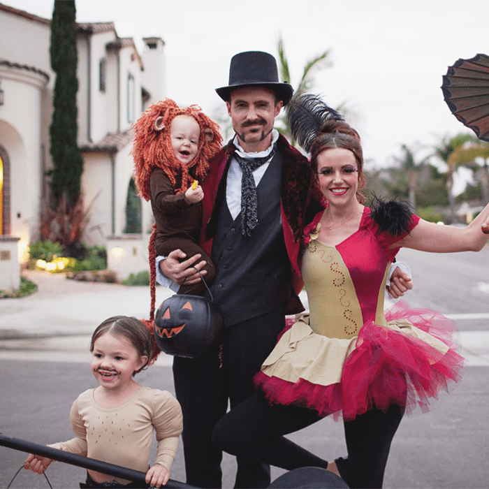 A family wearing circus-themed costumes poses together cheerfully on a residential street lined with houses and trees.