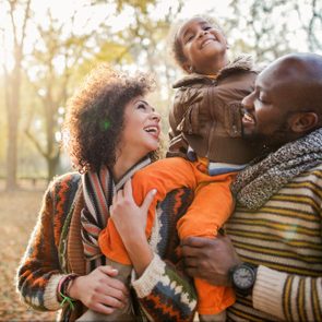 Child smiling, lifted by parents, surrounded by autumn trees.