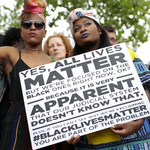 People hold protest signs emphasizing focus on Black lives amid others; outdoors amid a crowd under trees.