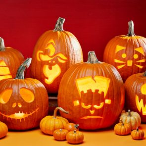 Pumpkins display carved jack-o'-lantern faces, glowing with light, surrounded by smaller pumpkins on a table, set against a red background.