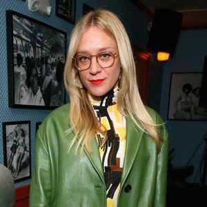 A person with long blonde hair wears glasses and a green coat, standing in a room decorated with framed photographs on the walls.