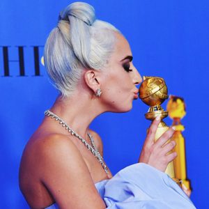 Person kisses golden award trophy, wearing blue gown and diamond jewelry, against a solid blue background.