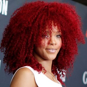Individual with vibrant curly red hair smiling, wearing a white outfit. Background is a dark backdrop with blurred white text partially visible.