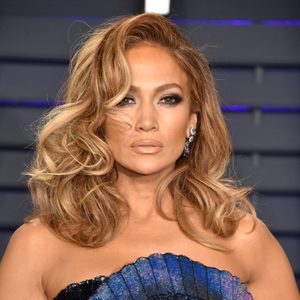 A person with styled wavy hair stands in front of a modern, dark backdrop, wearing blue and indigo attire.
