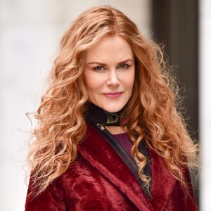Person with long, wavy hair stands outdoors, wearing a red coat and black scarf against a blurred urban background.