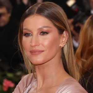 A person with long hair smiles slightly, wearing metallic ear jewelry, in a formal indoor event with blurred background elements.