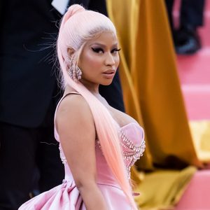 A woman poses confidently wearing a pink gown adorned with pearls. She stands in a formal event setting with a colorful backdrop.