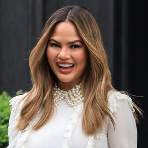 A person smiles broadly, wearing a white outfit with pearl details, set against a dark background with green foliage.