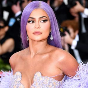 Person with long purple hair looks sideways, wearing a sparkling outfit, surrounded by people in a bright, formal event setting.