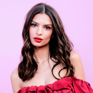 Person posing against a pink background, wearing a red off-the-shoulder dress, with wavy brown hair and red lipstick.