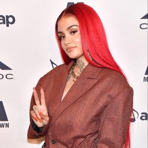 Person with red hair shows peace sign, wearing a brown blazer against a backdrop with logos.