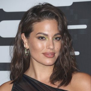 A woman smiles subtly, wearing green eyeshadow and layered earrings, set against a dark patterned background.