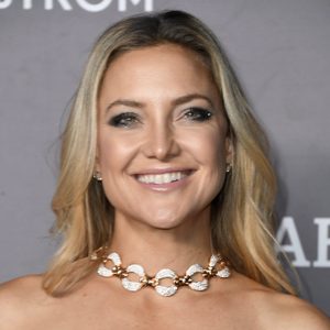 A person smiles while wearing a decorative necklace; standing against a neutral gray background.
