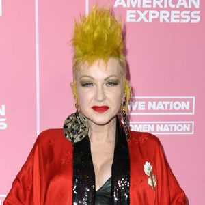 Person poses; wearing large earrings, red jacket, black top; standing against a pink background with 