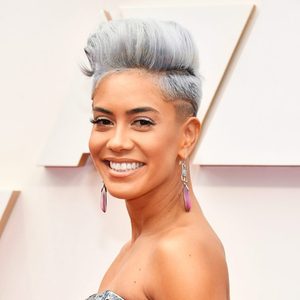 A person smiles with short, styled gray hair, wearing long pink earrings against a simple, light-colored background.