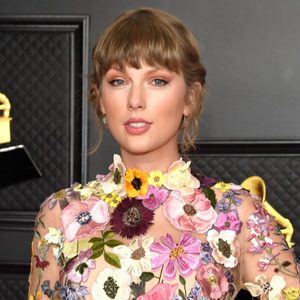 A person stands wearing a floral-embroidered dress, posing gracefully on a black-carpeted stage with a golden award visible in the background.