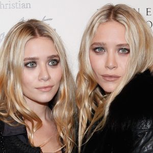 Two women stand closely, looking at the camera, surrounded by a neutral backdrop with partial text in the background.