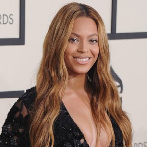 A woman smiles, wearing a black dress with long hair. The background shows an awards event with framed logos and a light-colored wall.