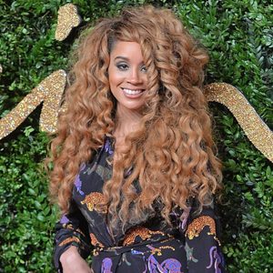Person with voluminous curly hair smiles, wearing a patterned dress. They stand against a green leafy backdrop with glittering gold details.
