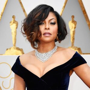 A woman poses in an elegant dress and diamond necklace, with golden statuettes and a white background behind her.