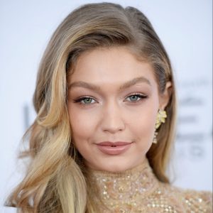 Person smiling, wearing a high-neck embellished outfit, with wavy blonde hair, and gold earrings. The background is plain blue-grey.