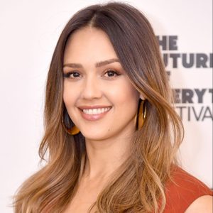 A person smiles facing forward, wearing large hoop earrings, in front of a backdrop with partially visible text on a white background.