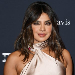 A person stands smiling, wearing a satin dress, with a backdrop featuring the text 