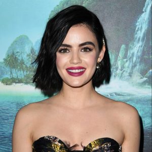 A person with dark hair smiles, standing in front of a tropical beach and waterfall backdrop.